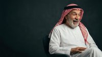 Middle-aged man with gray beard wearing traditional white thobe and red checkered ghutra headwear, holding an open book against dark background. Suitable for educational, cultural, and business content.