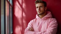 Young Caucasian male model wearing pink hoodie standing by window with crossed arms. Dramatic red wall and window shadow creates modern fashion portrait lighting.
