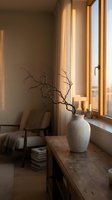 Minimalist interior with ceramic vase and dried branches on wooden table near window with warm sunlight creating cozy atmosphere for home decor.