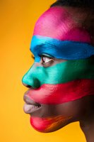 Profile portrait of African woman with vibrant multicolored face paint stripes in pink, blue, green and red against yellow background, showcasing creative makeup artistry.