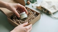 Caucasian woman's hands placing a small glass candle jar into an eco-friendly kraft box with shredded paper filler for sustainable gift packaging.