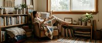 Young woman relaxing with book in cozy living room with wooden furniture, bookshelves and natural light streaming through windows for lifestyle content.