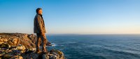 Middle-aged man with reddish-brown hair, Caucasian ethnicity, wearing casual outdoor jacket standing on rocky cliff overlooking vast ocean horizon at sunset. Contemplative traveler gazing at seascape.