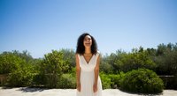 Young woman with long dark curly hair and olive skin tone wearing white sleeveless dress standing in sunny garden with green shrubs and clear blue sky, suitable for lifestyle and wellness content.