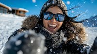 Young African American woman laughing while taking selfie on winter vacation, wearing sunglasses and warm clothing against snowy mountain backdrop with flying snow.