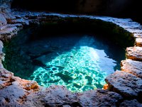 Natural cave pool with crystal clear turquoise water reflecting sunlight through rocky limestone formation, creating ethereal underwater glow and ripple patterns.