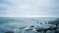 Serene coastal landscape with smooth stones on beach and calm ocean waves under overcast sky creating peaceful atmosphere for relaxation and meditation.