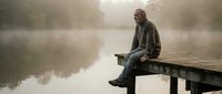 Contemplative senior man sitting on wooden dock by misty lake during peaceful morning, reflecting solitude and tranquility in natural outdoor setting.