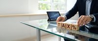 Man in business suit arranging wooden blocks spelling tax percent symbol at glass desk with laptop displaying financial graph in modern office with window.