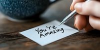 Hand writing positive affirmation message on white paper note with metallic pen against rustic wooden table surface in soft natural lighting.