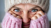 Close-up portrait of senior Caucasian woman with bright blue eyes wearing knitted beige hat and pink sweater, covering part of her face with hands during winter.