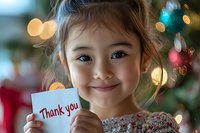 Young Asian girl holding thank you note card with sweet smile, colorful bokeh lights and Christmas decorations creating warm holiday atmosphere in background.