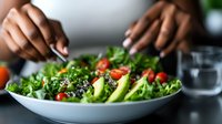 Fresh green salad with avocado slices, cherry tomatoes and sesame seeds being prepared by African American woman hands. Healthy eating and meal preparation.