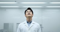 Young Asian male scientist or laboratory technician wearing white lab coat and glasses, looking upward thoughtfully in modern sterile laboratory setting. Suitable for healthcare, research, and medical education content.