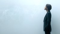 Young Asian woman in black business suit looking up at foggy sky with contemplative expression, side view against minimalist misty background.