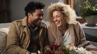 Young African American couple sharing genuine laughter while relaxing on couch with fresh flowers, expressing joy and authentic relationship moments in casual home setting.