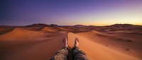 Traveler's boots extended over golden sand dunes at sunset, showcasing desert adventure and wanderlust exploration with vast landscape views.