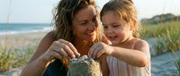 Adult woman with long brown hair and young child with reddish-blonde hair building sandcastle together on sandy beach at sunset, ocean and dune grass visible in background, summer family vacation moment.