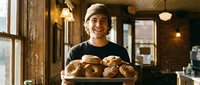 Male baker holding fresh baked bread rolls in rustic bakery shop interior with warm lighting and wooden details for food business and artisan bakery concepts.