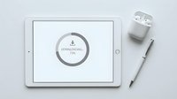 Tablet displaying a downloading progress circle at 73%, placed beside wireless earbuds in a case and a stylus pen on a white surface.