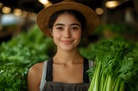 Young Asian woman farmer in straw hat and overalls holding fresh celery stalks, surrounded by organic produce in natural lighting at local market.