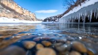 Frozen lake with dramatic icicles hanging from rocky cliffs, smooth pebbles visible underwater, winter landscape with snow-covered mountains and clear blue sky.