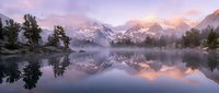 Serene mountain lake at sunrise with perfect reflections of snow-capped peaks and pine trees in calm water under pastel pink and purple sky.