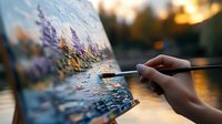 Artist hand holding paintbrush creating colorful landscape painting on canvas outdoors during golden hour sunset with warm natural lighting for creative art projects.