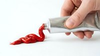 Hand squeezing red paint from a metal tube onto a white surface, creating a vivid swirl of crimson color for art and design concepts.