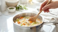 Hand stirring homemade chicken noodle soup with vegetables in pot on stove. Comfort food cooking preparation in modern kitchen with wooden spoon.