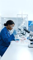 Female scientist of South Asian descent wearing safety glasses and blue lab coat conducting microbiological research with petri dishes and microscopes in modern laboratory setting for healthcare and scientific studies.