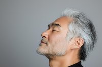 Profile of mature Asian man with gray hair and beard in black turtleneck, eyes closed with serene expression against gray background, showing mindfulness and peace.