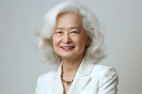 Senior Asian woman with silver hair wearing white blazer smiles warmly against neutral background, expressing confidence and joy in professional portrait setting.