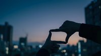 Hands forming frame gesture against blurred city skyline at dusk with bokeh lights creating artistic composition for creative vision concepts.