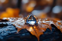 Sparkling blue crystal ring on autumn maple leaf covered with water drops against dark background with golden bokeh lights and reflections.