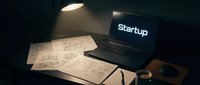 Laptop with startup text on screen, architectural blueprints, coffee cup on dark desk under lamp light, entrepreneurship and business launch concept.