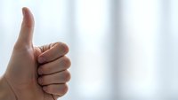 Hand showing thumbs up gesture against bright window background expressing approval and positive feedback in modern interior space.