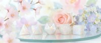 Elegant white geometric candles and holders arranged on glass shelf with soft pastel flowers including roses and hydrangeas in background for home decor and events.