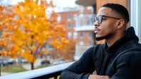 Young African American man in glasses and black hoodie looking thoughtfully from balcony with autumn foliage and urban residential building in background.