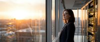 Professional Asian businesswoman in elegant black blazer standing by office window during golden hour sunset, contemplating city skyline in modern corporate building.