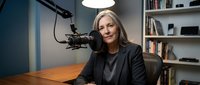Professional mature blonde woman podcaster speaking into microphone at home studio desk with bookshelf background for broadcasting content creation.