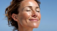 Middle-aged woman with closed eyes enjoying sunshine outdoors, expressing serenity and contentment with a gentle smile against clear blue sky background.