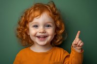 Happy redhead toddler girl with curly hair pointing finger up showing excitement and joy against green background for educational content.