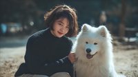 Young Asian woman with short hair in black sweater embracing fluffy white Samoyed dog outdoors in warm sunlight, creating heartwarming pet companionship moment.