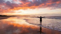 Person with arms outstretched celebrating freedom on beach at golden sunset with dramatic clouds reflecting in wet sand and ocean waves.