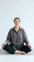 Young Caucasian woman practicing mindfulness meditation in lotus position with eyes closed, wearing casual gray linen shirt on yoga mat for wellness and relaxation.