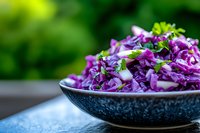 Fresh purple cabbage coleslaw with herbs and onion in dark ceramic bowl on blue surface against blurred green background. Healthy vegetarian salad.