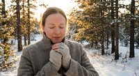 Young woman enjoying peaceful moment in snowy winter forest wearing warm gray coat and mittens with eyes closed and serene expression during golden hour.