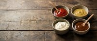 Four ceramic bowls with different condiments and sauces including ketchup, mustard, mayonnaise and barbecue sauce on rustic wooden table background.