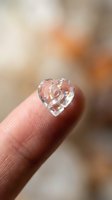 Tiny transparent crystal heart held delicately on fingertip with warm bokeh background for love romance jewelry crafts miniature collectibles.